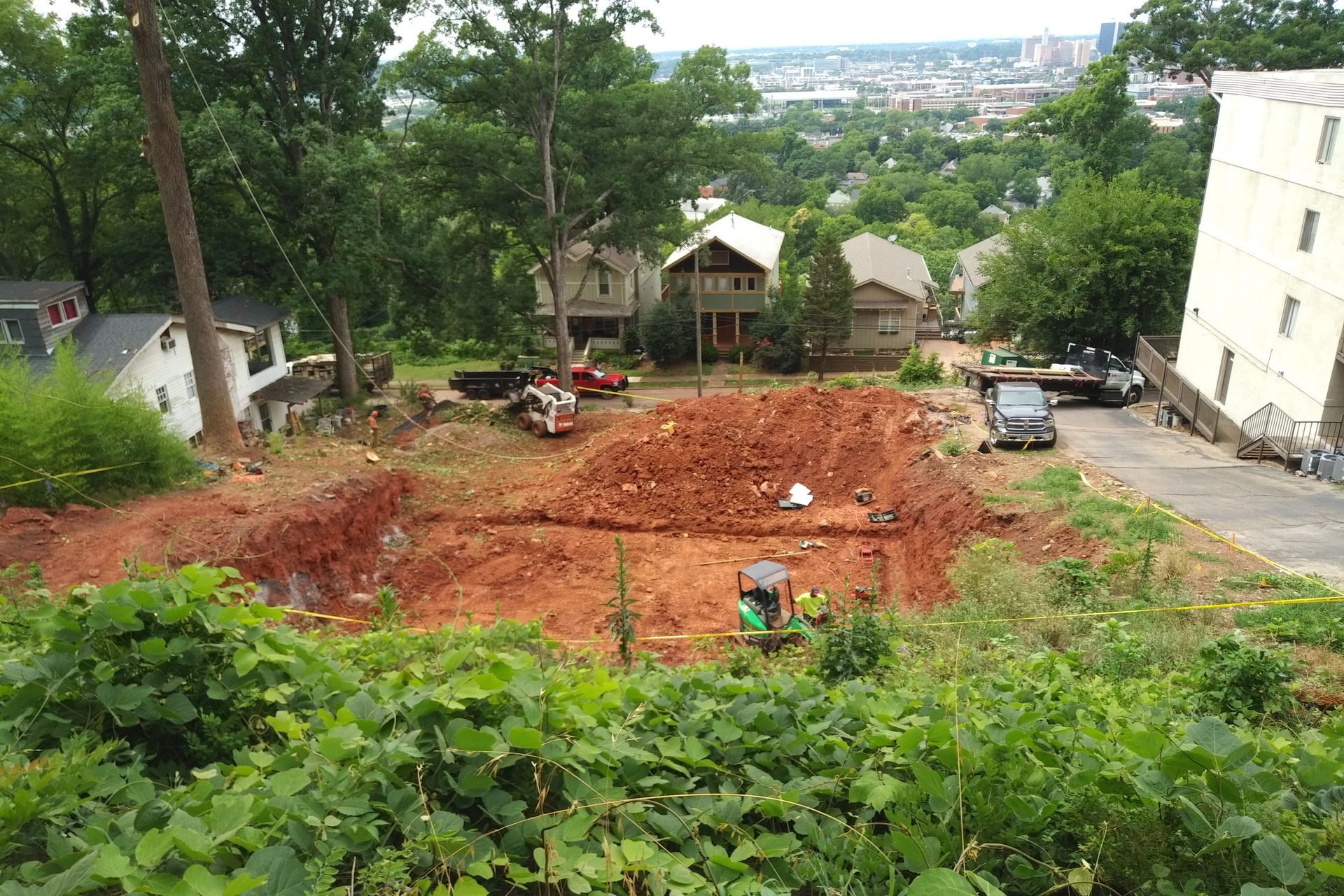 Construction site on a hillside with red dirt, excavators, and a view of houses and the city.