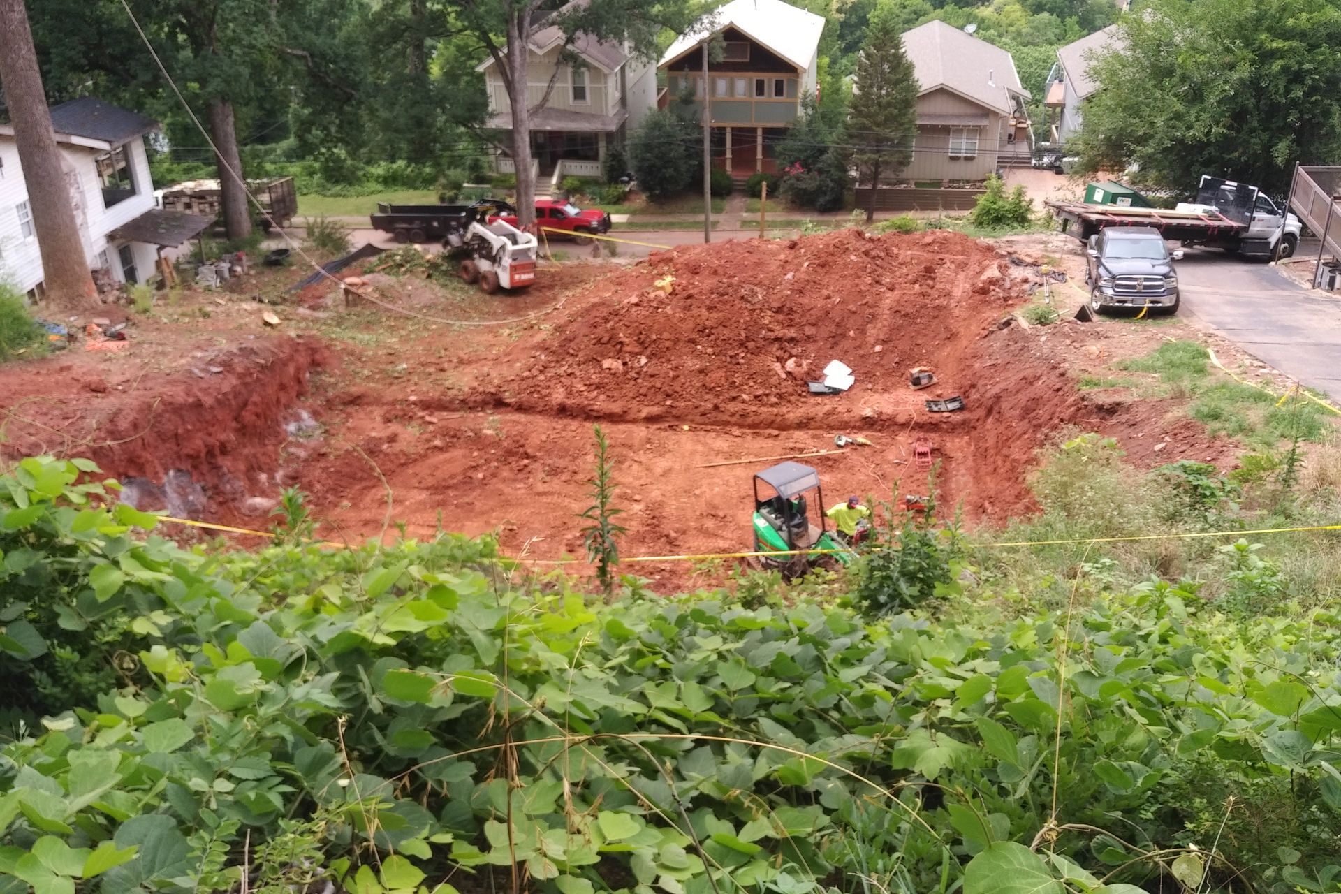 Construction site with red soil, excavation, equipment, and houses in the background.