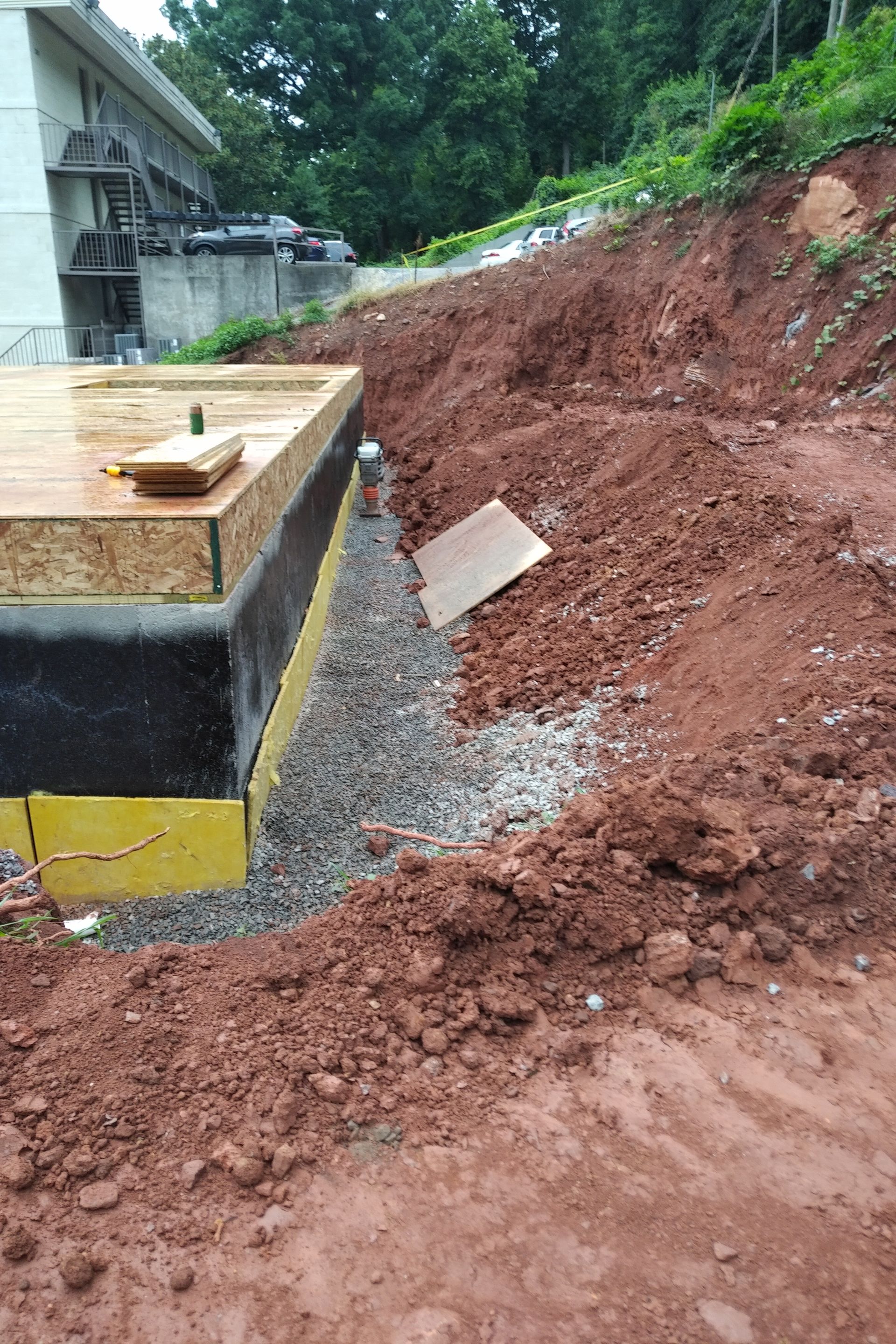 Construction site with gravel drainage against a building foundation and sloped red dirt.