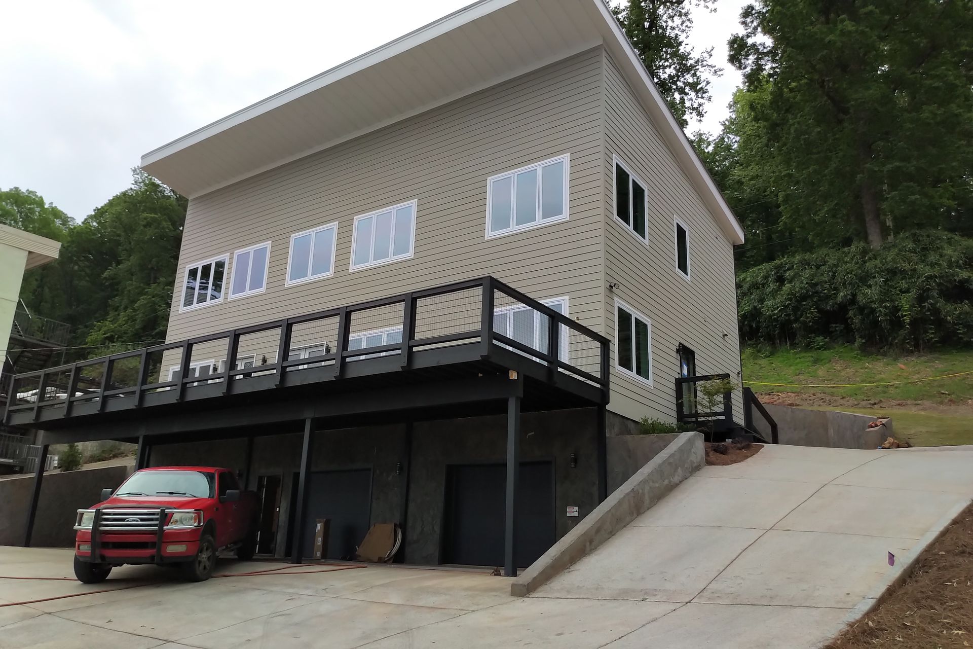 Beige house with black deck, parked red truck, concrete driveway, and green hill backdrop.