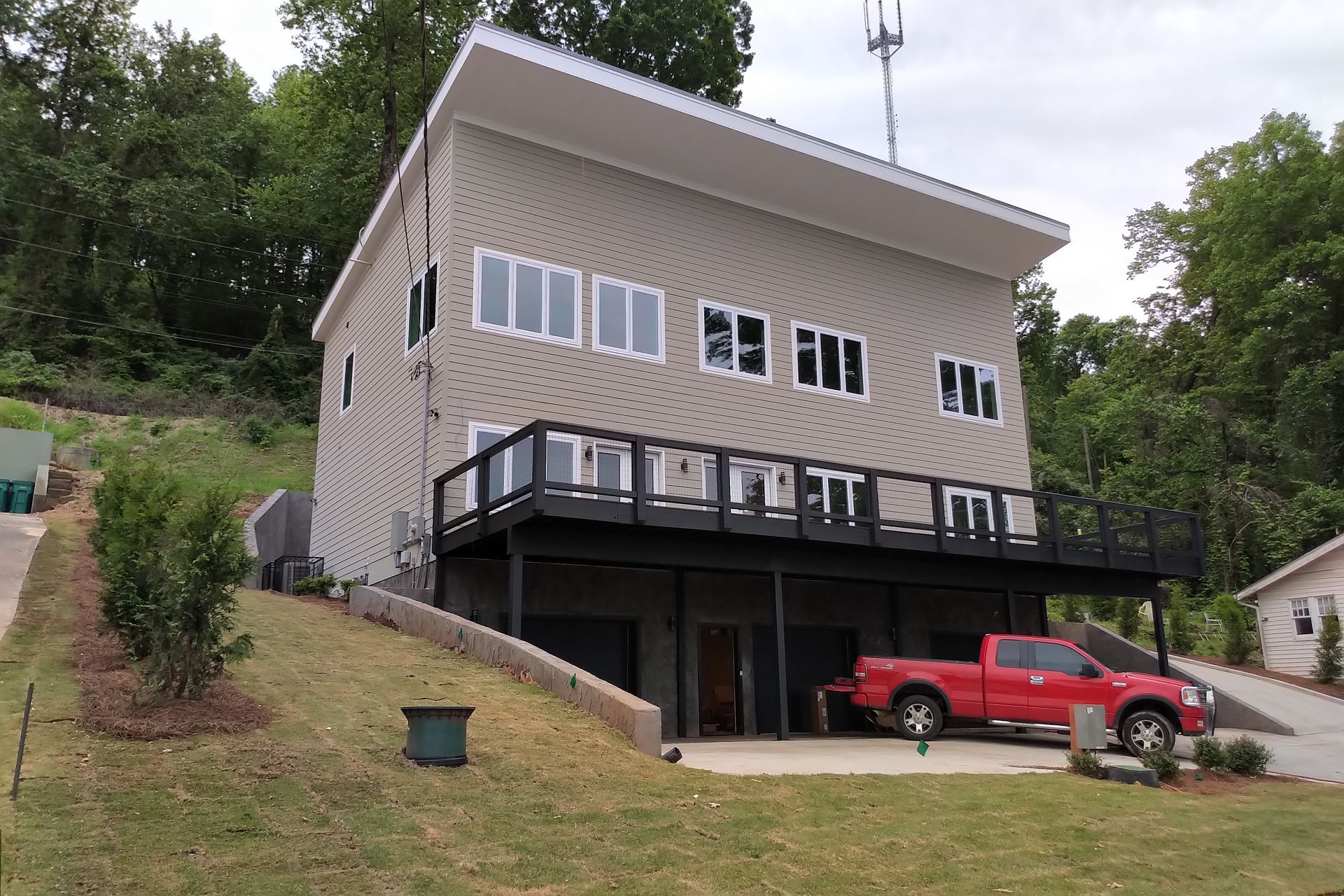 Two-story house with red truck parked underneath. Tan siding, black deck, set on hillside.