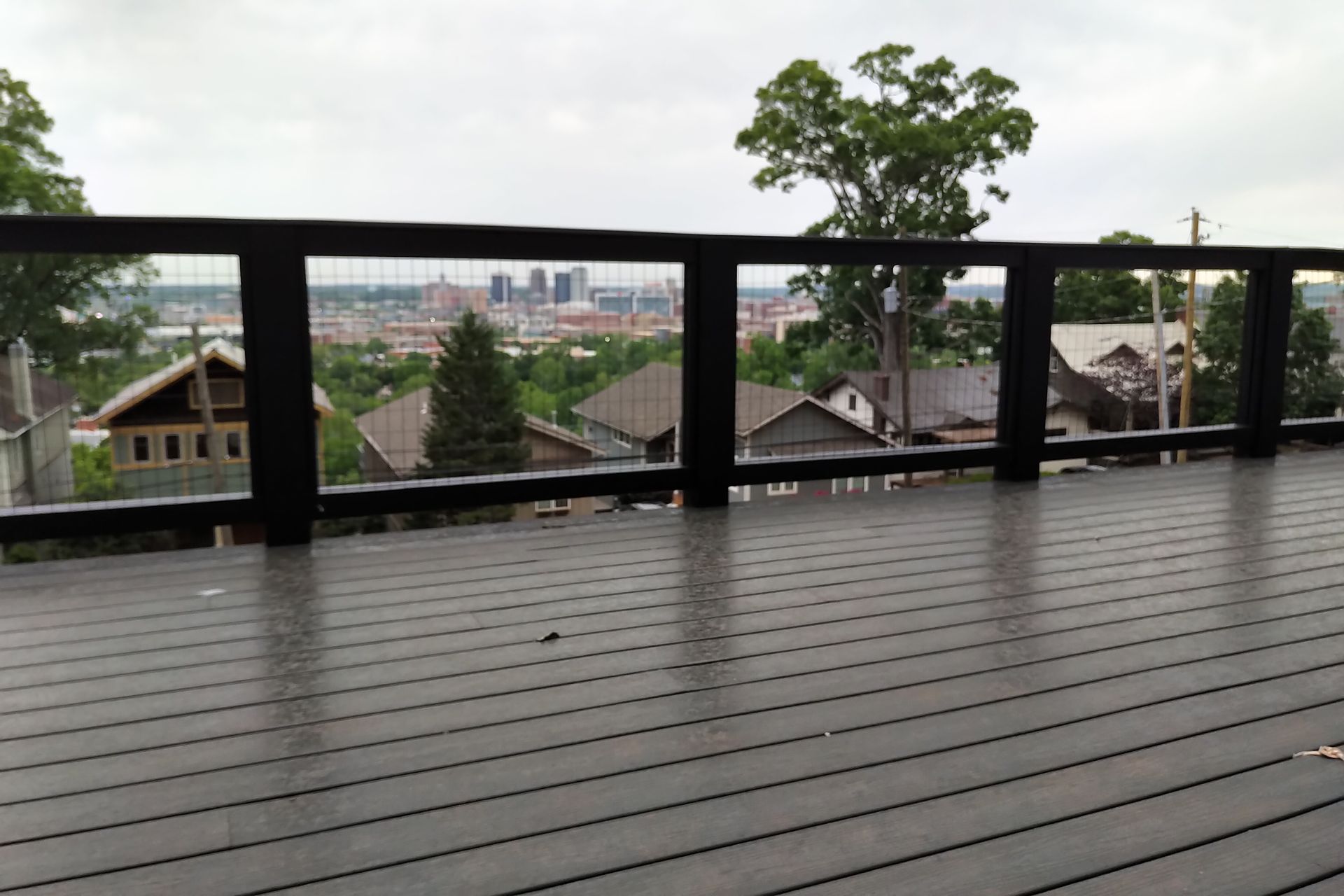Deck overlooking a city skyline. Gray deck, black railing, and cloudy sky.