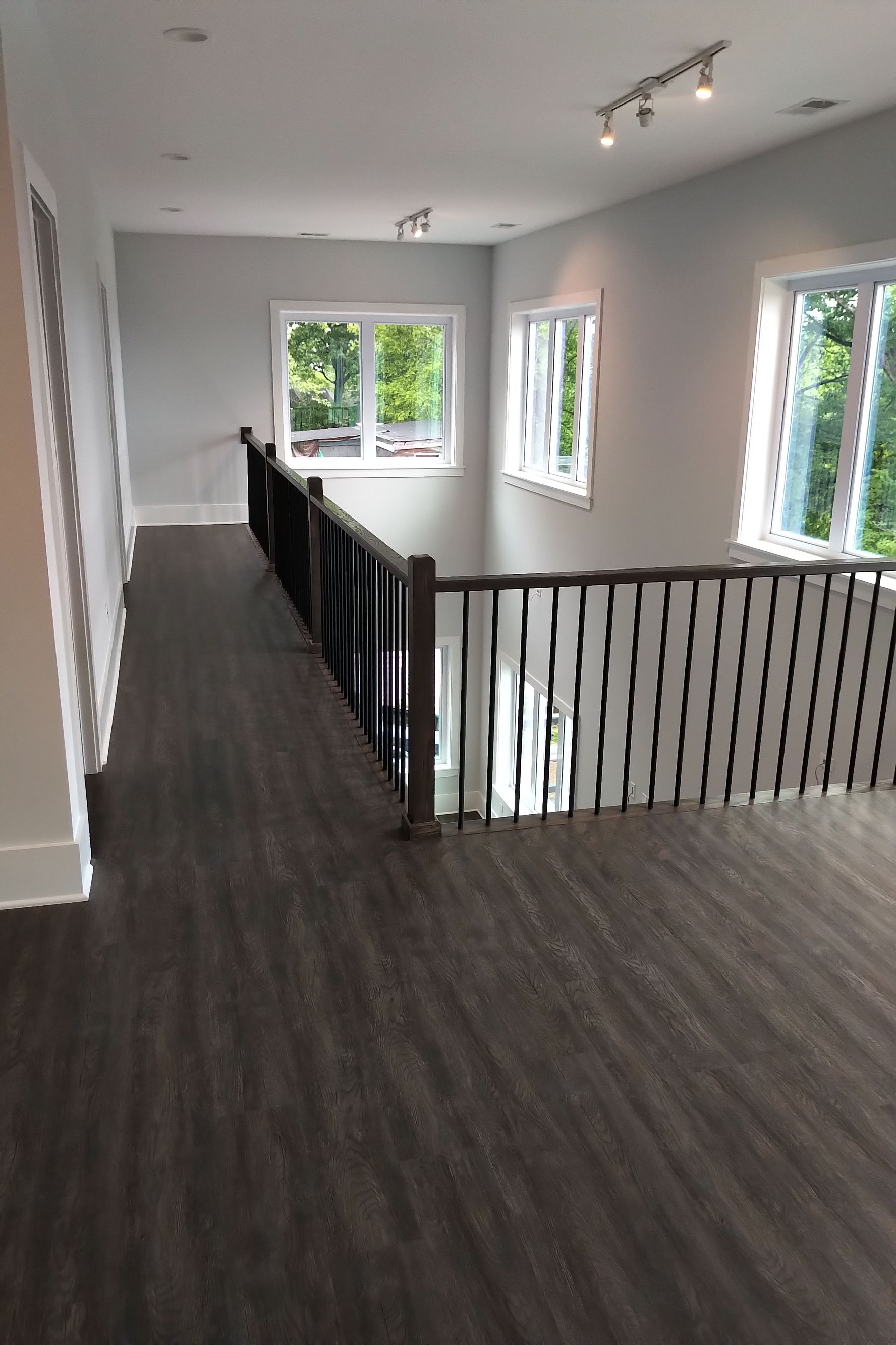 Dark wood-floored hallway with black railing overlooking a lower level. Light gray walls, three windows.