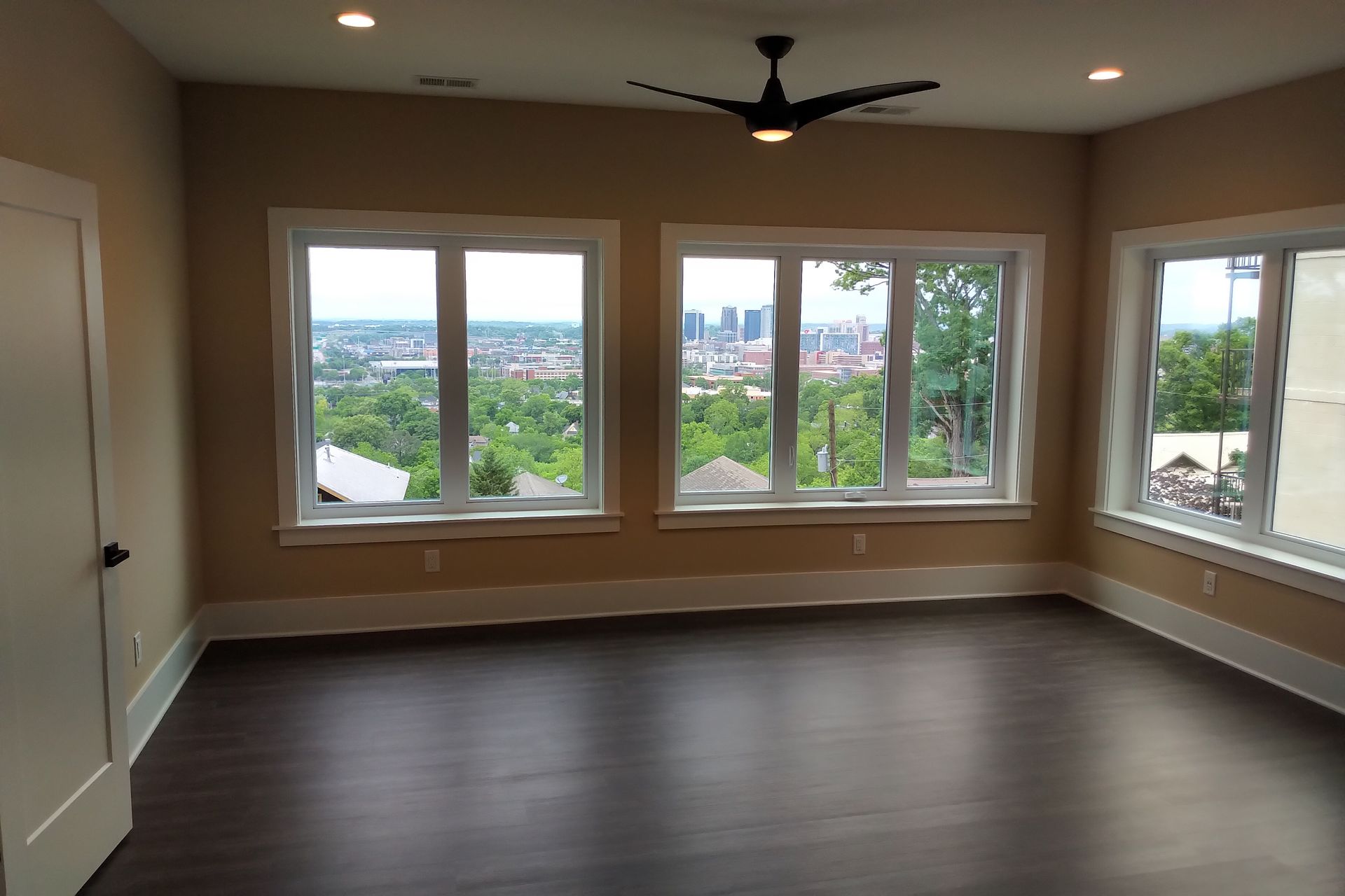 Empty room with large windows overlooking a city. Dark wood floors, light walls, and a ceiling fan.