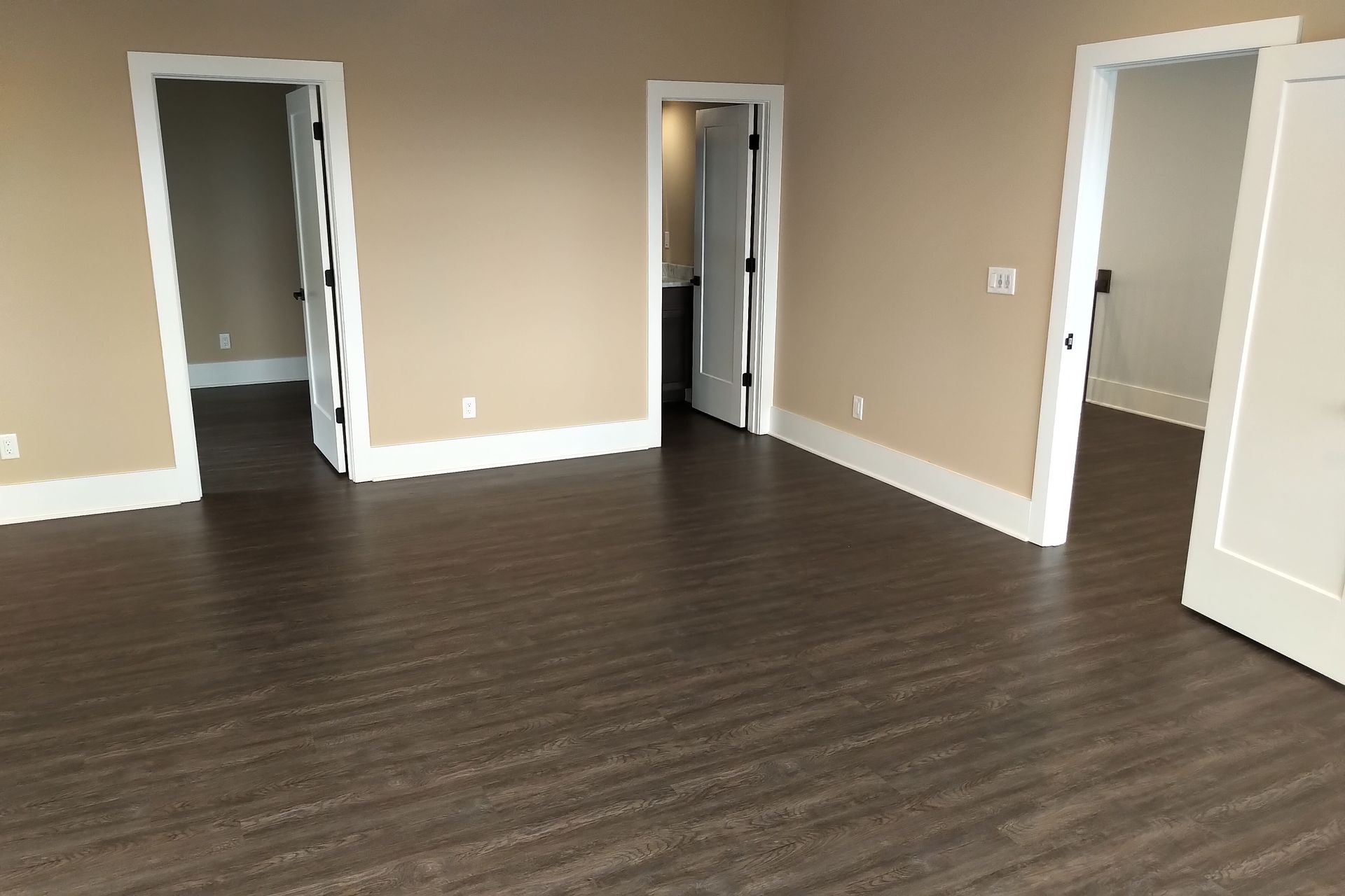 Empty room with brown flooring and beige walls. Three white-framed doorways lead to other rooms.