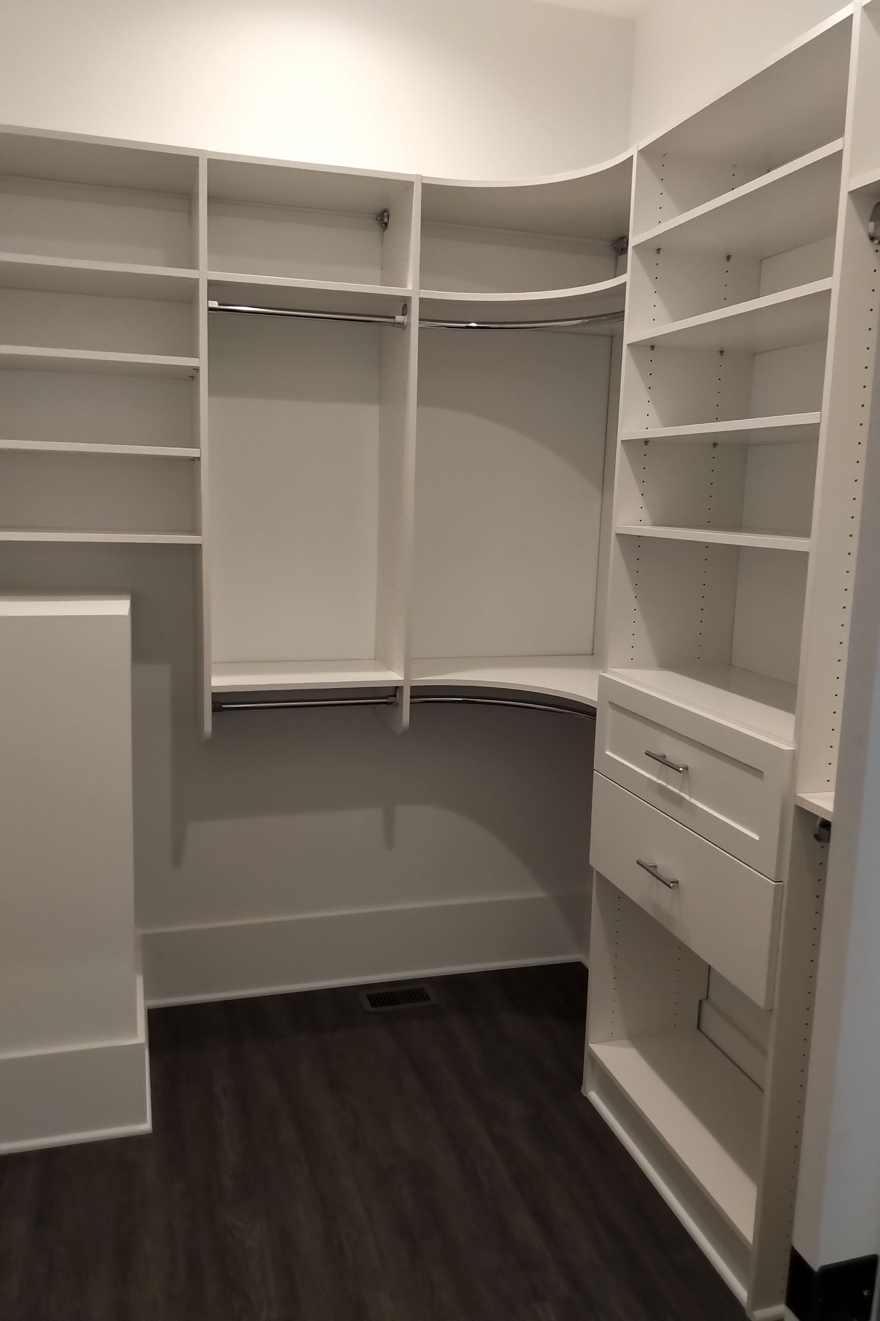 White walk-in closet with shelves, drawers, and hanging rods. Dark wood floor, white walls.