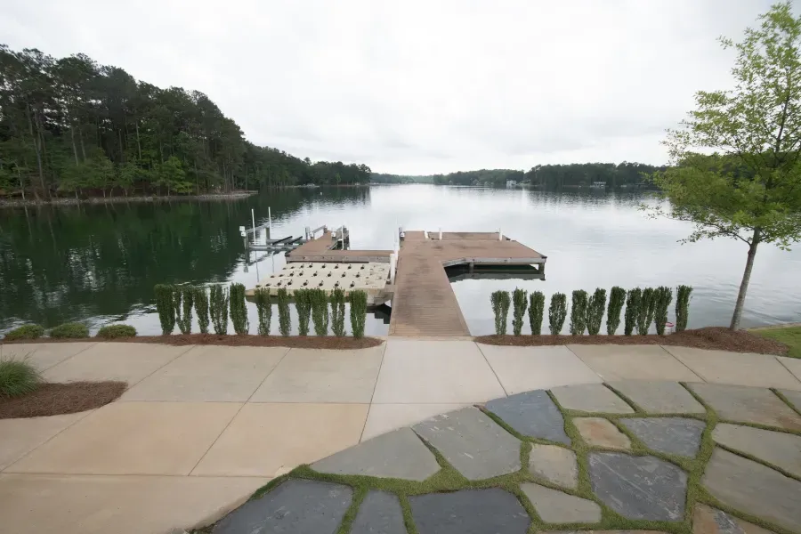 Lakefront view with wooden dock, concrete pathway, and green hedges. Cloudy day with trees in the background.