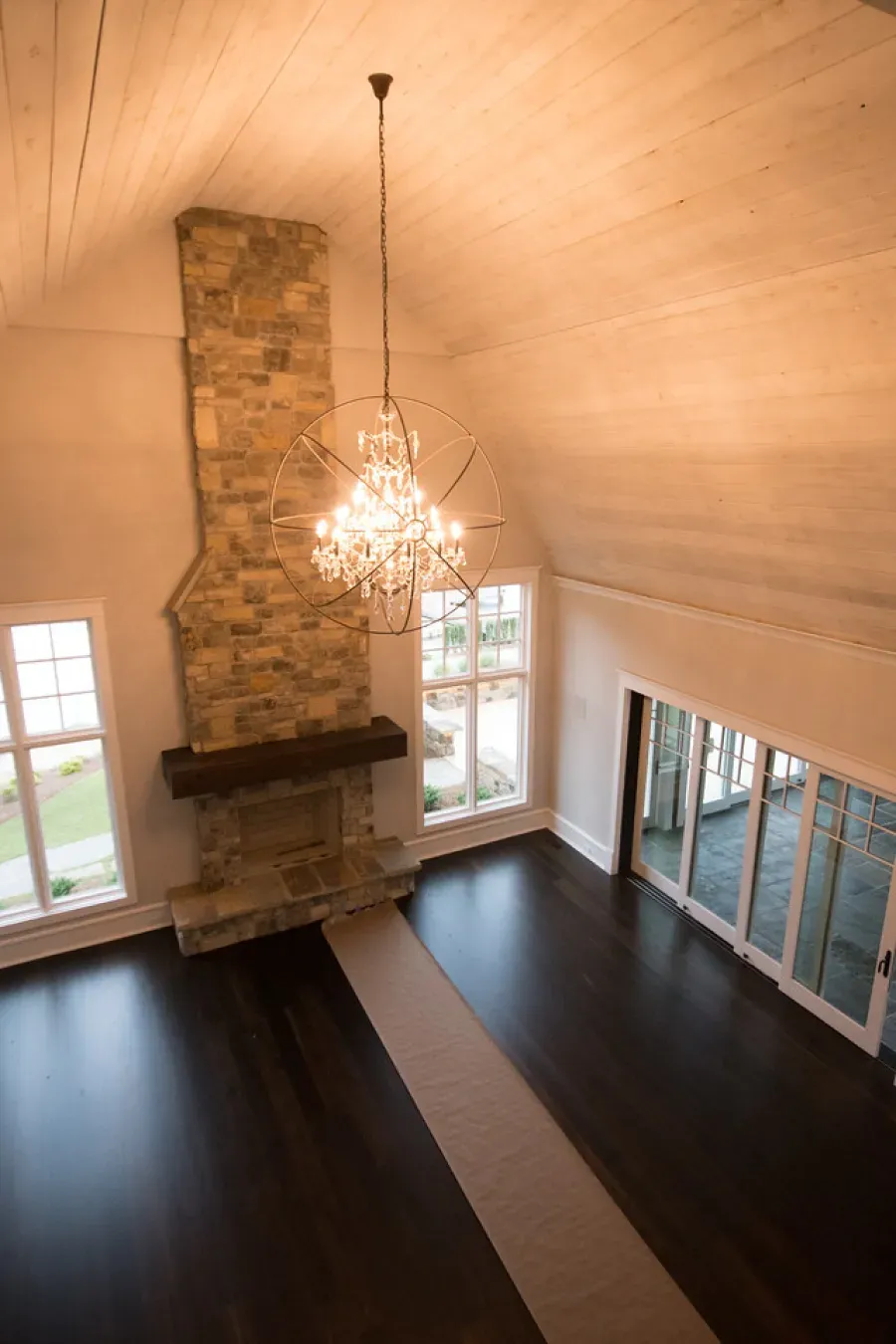 High-angle view of a room with a stone fireplace, chandelier, dark wood floor, and large windows.