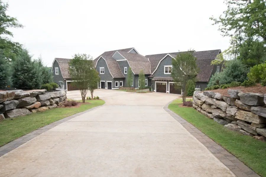 Driveway leading to a gray house with gabled roofs, surrounded by trees and rock walls.