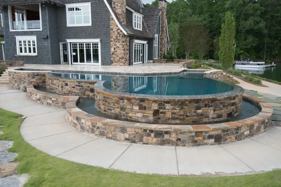 Stone-edged pool with cascading water, overlooking a lake, beside a gray house.