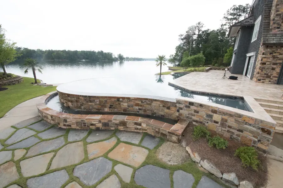 Stone patio and pool overlooking a lake, next to a house with trees in the background.