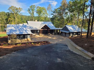 A rustic house with a white roof and a gravel driveway surrounded by trees and greenery.