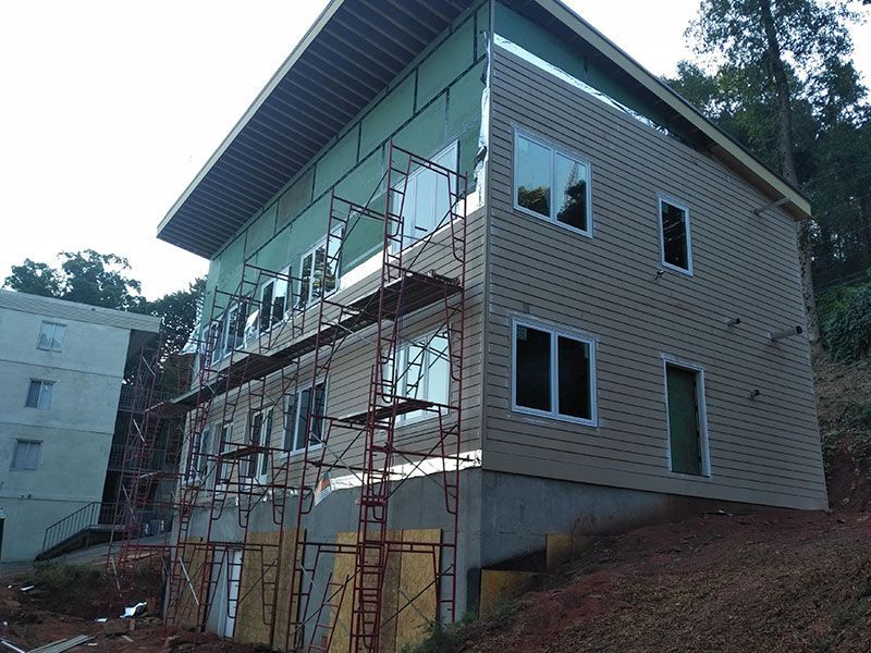 Three-story building under construction with tan siding, scaffolding, windows, and a hillside setting.