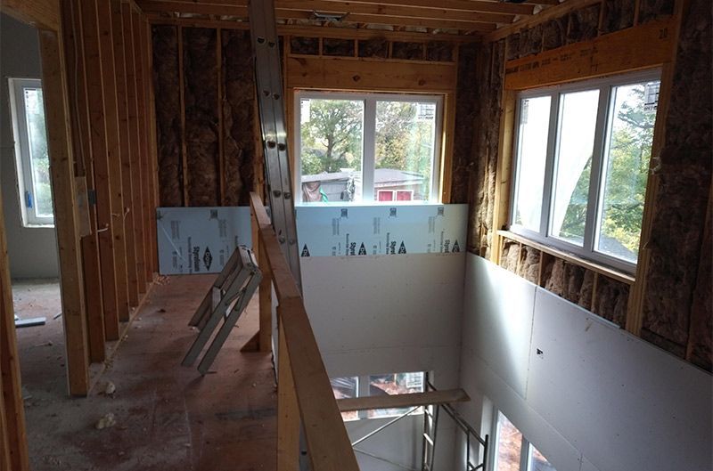 Interior view of a house under construction. Wood framing, insulation, windows, and a staircase are visible.