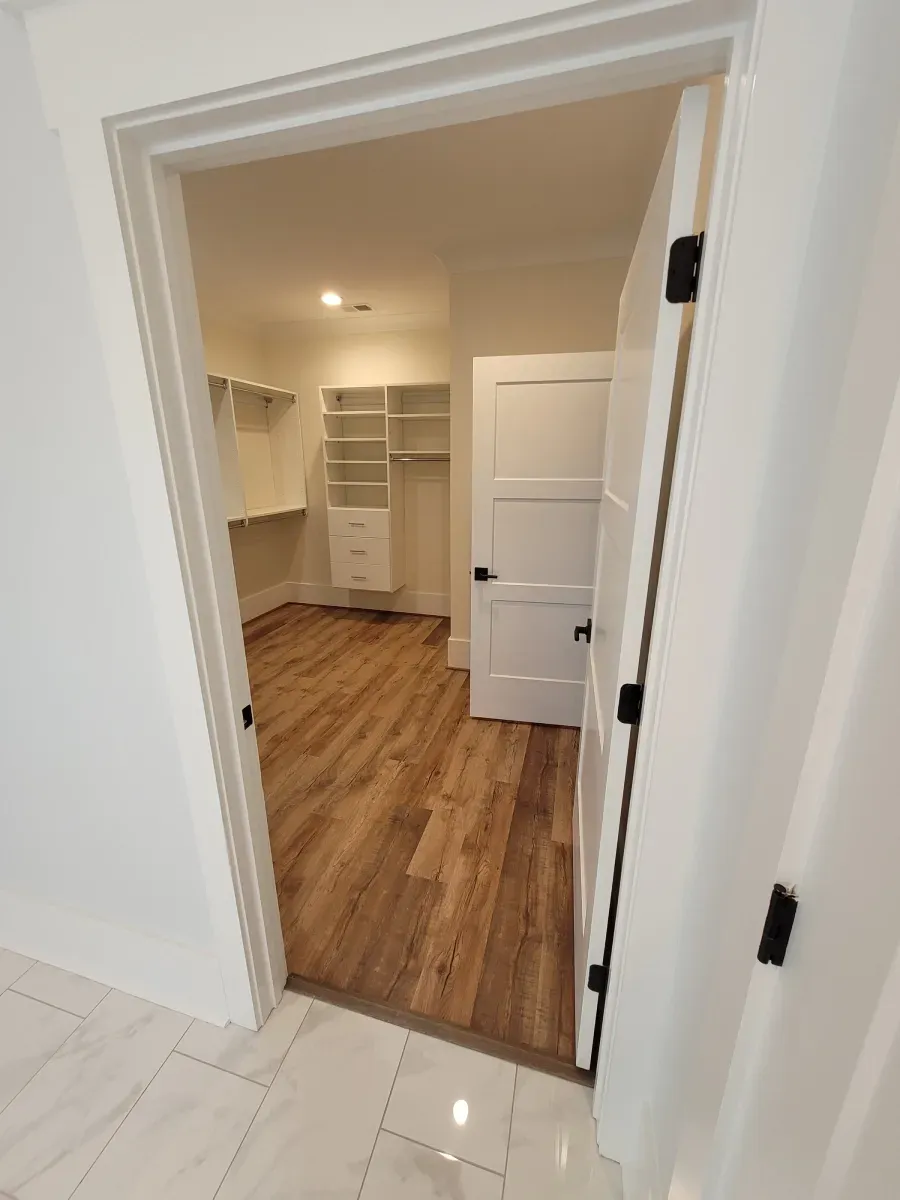 Open doorway to a walk-in closet with white shelves, cabinets, and door. Wood floors and white walls.