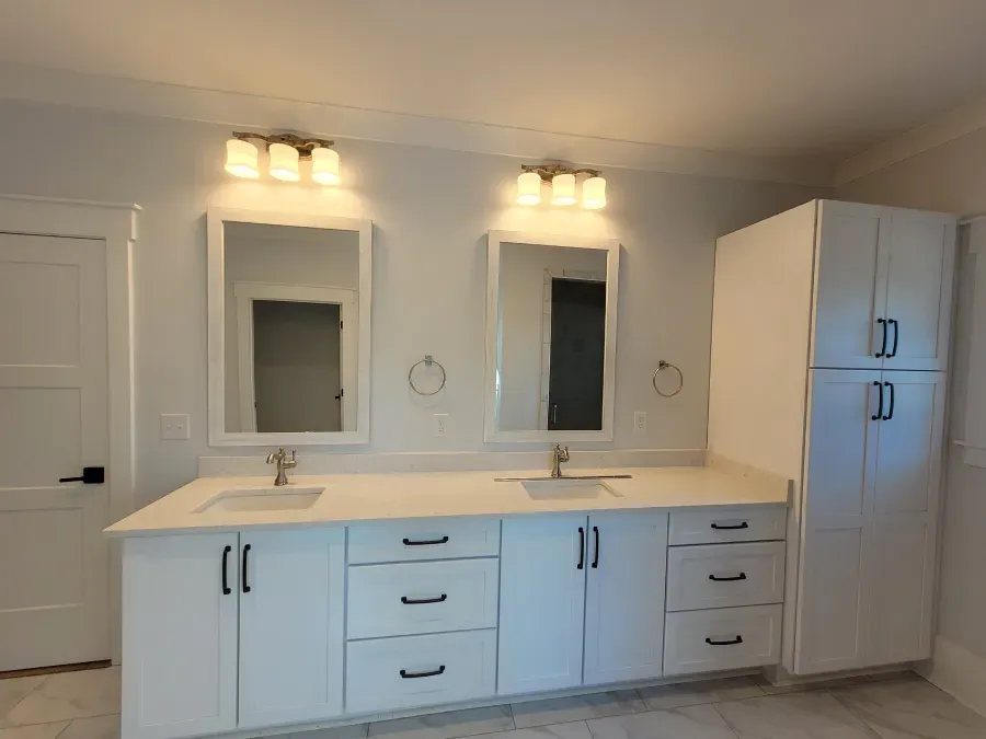White bathroom with double vanity, mirrors, and storage cabinet. Two sinks, gold fixtures, and dark hardware.