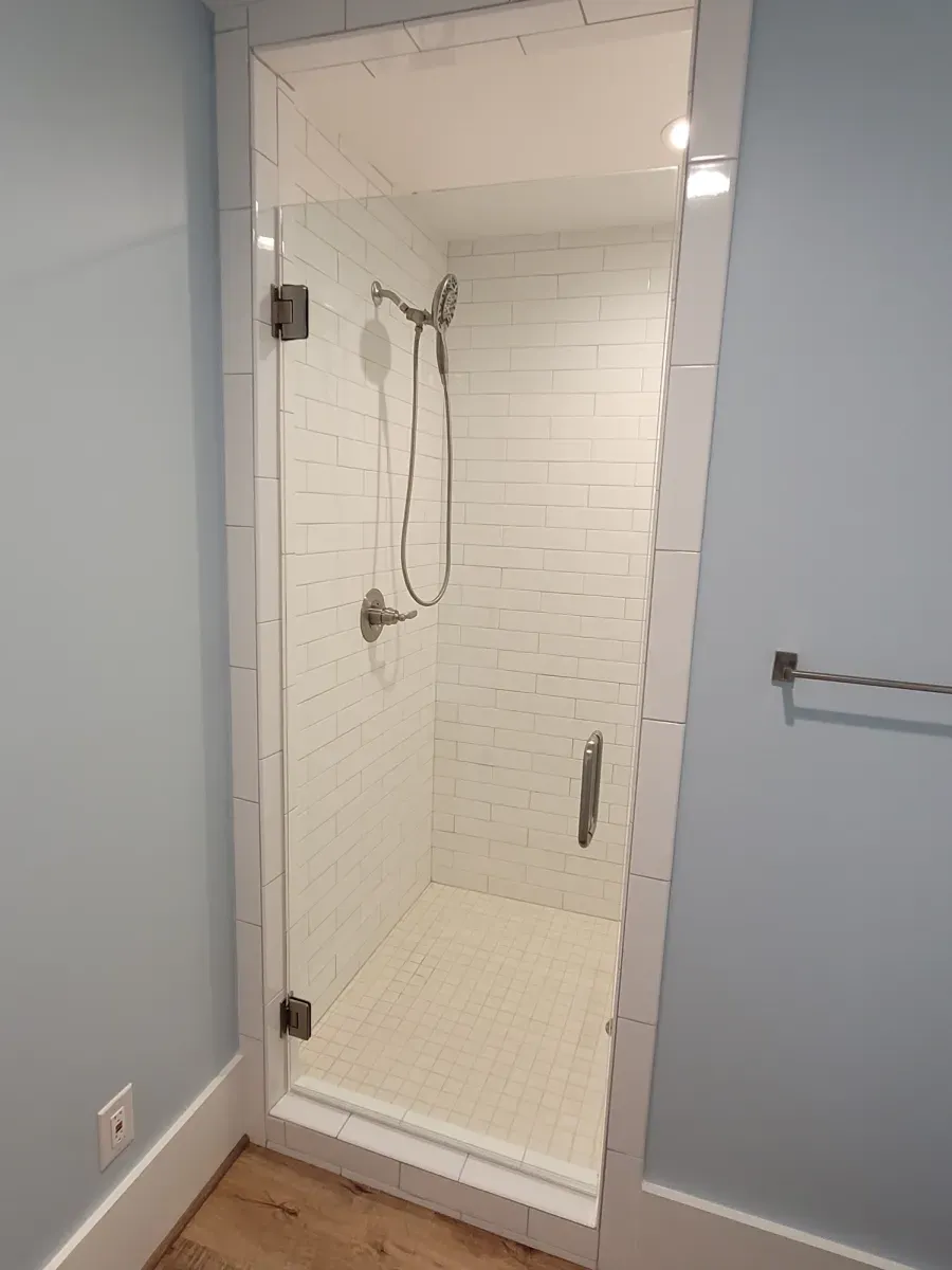 A glass-door shower with white tile walls, inside a room with light-blue walls and a wood floor.