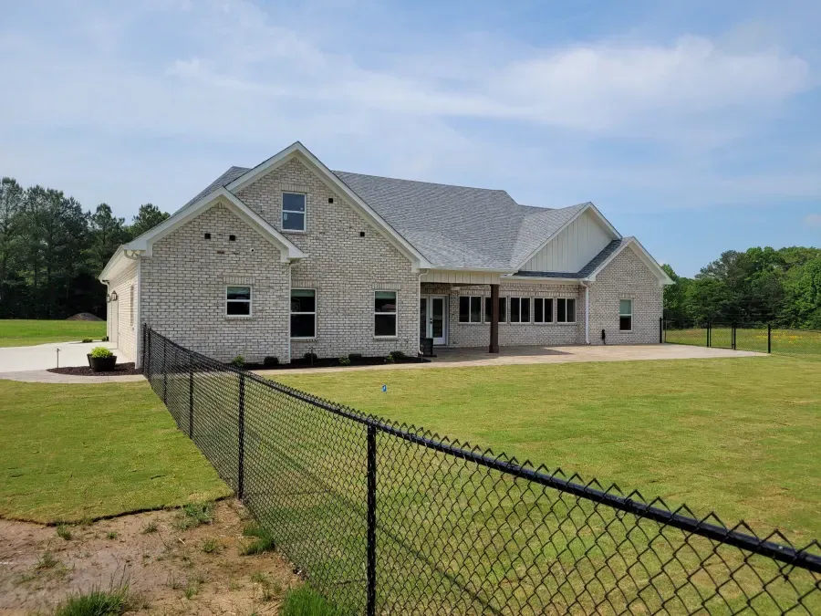 Brick house with gray roof, black chain-link fence, and large green lawn under a blue sky.