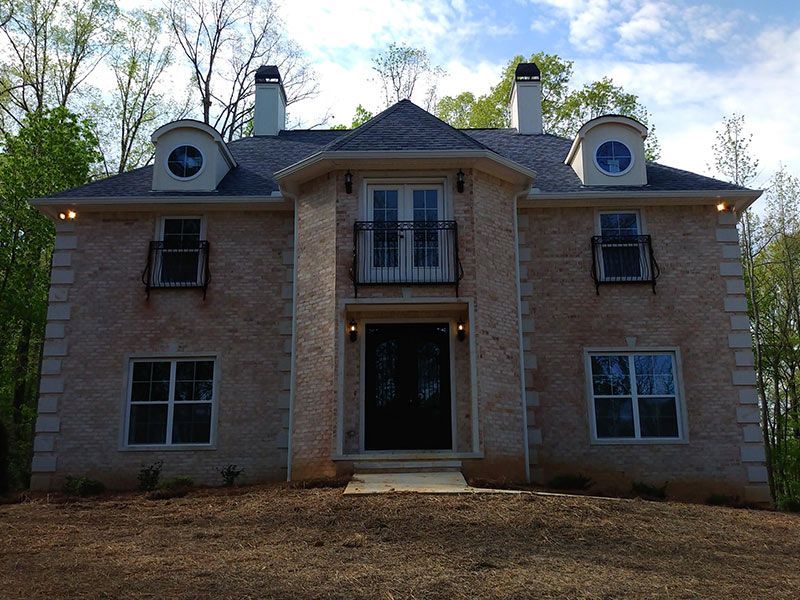 Two-story beige house with black trim and balconies, set against a wooded backdrop under a cloudy sky.