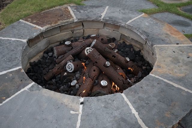 Fire pit with decorative logs and flames, surrounded by stone pavers and a stone border.
