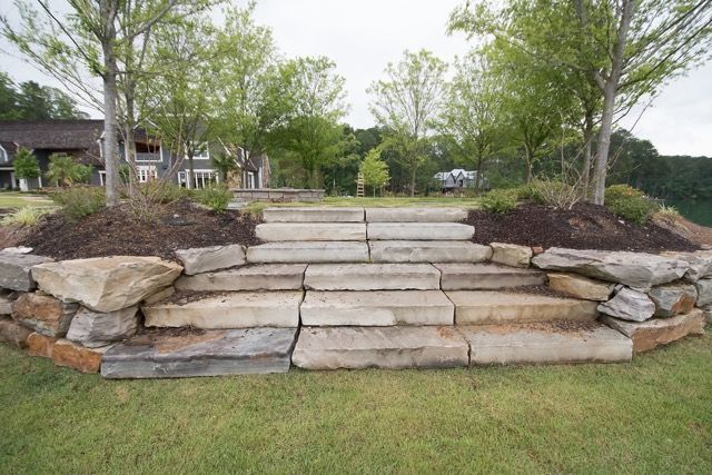 Stone steps leading to a higher level, flanked by stone walls and young trees, in a grassy area.