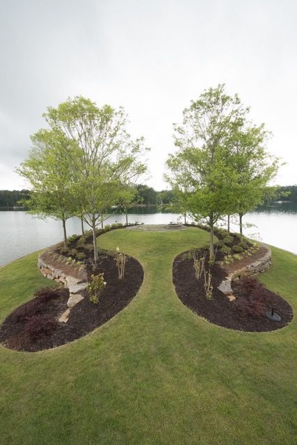 Two trees on a small peninsula with curving flower beds, grass, and a lake in the background.