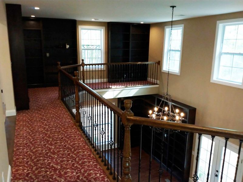 Second-floor hallway with red carpet, railing, bookshelves, and windows overlooking a chandelier and lower level.