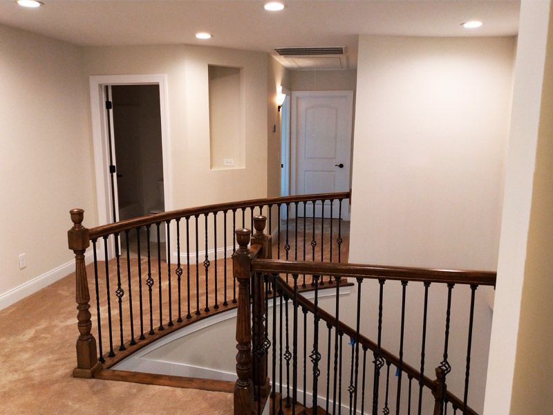 Interior view of a curved wooden staircase with ornate metal railings leading to a hallway with doors and recessed lighting.
