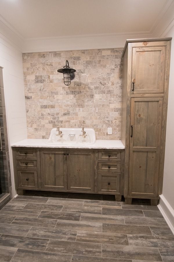 Bathroom with a rustic wood vanity and cabinet, brick-style backsplash, marble countertop, and gray tile floor.
