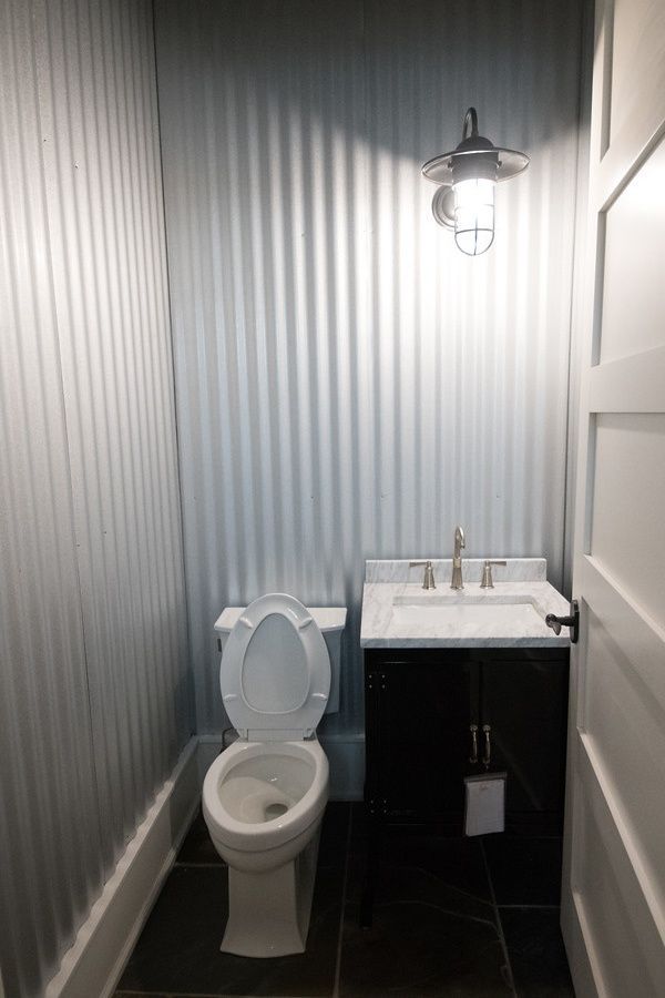 Small bathroom with corrugated metal walls, toilet, sink, and black cabinet; white door.