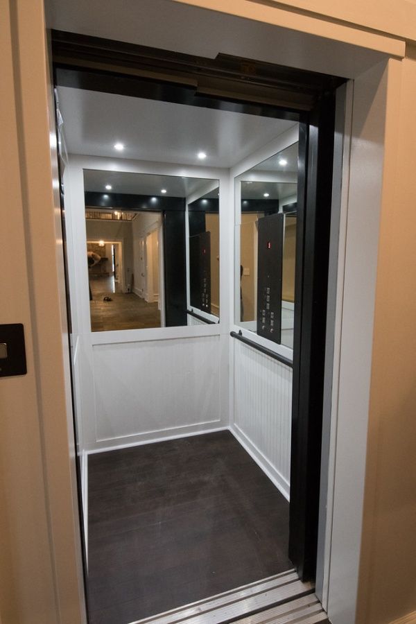 Interior view of a small elevator with white paneling, mirrors, and dark floors, framed by a black door.