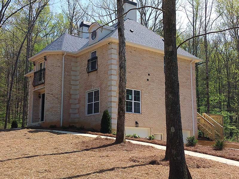 Two-story beige brick house with black balconies, surrounded by trees, on a sunny day.