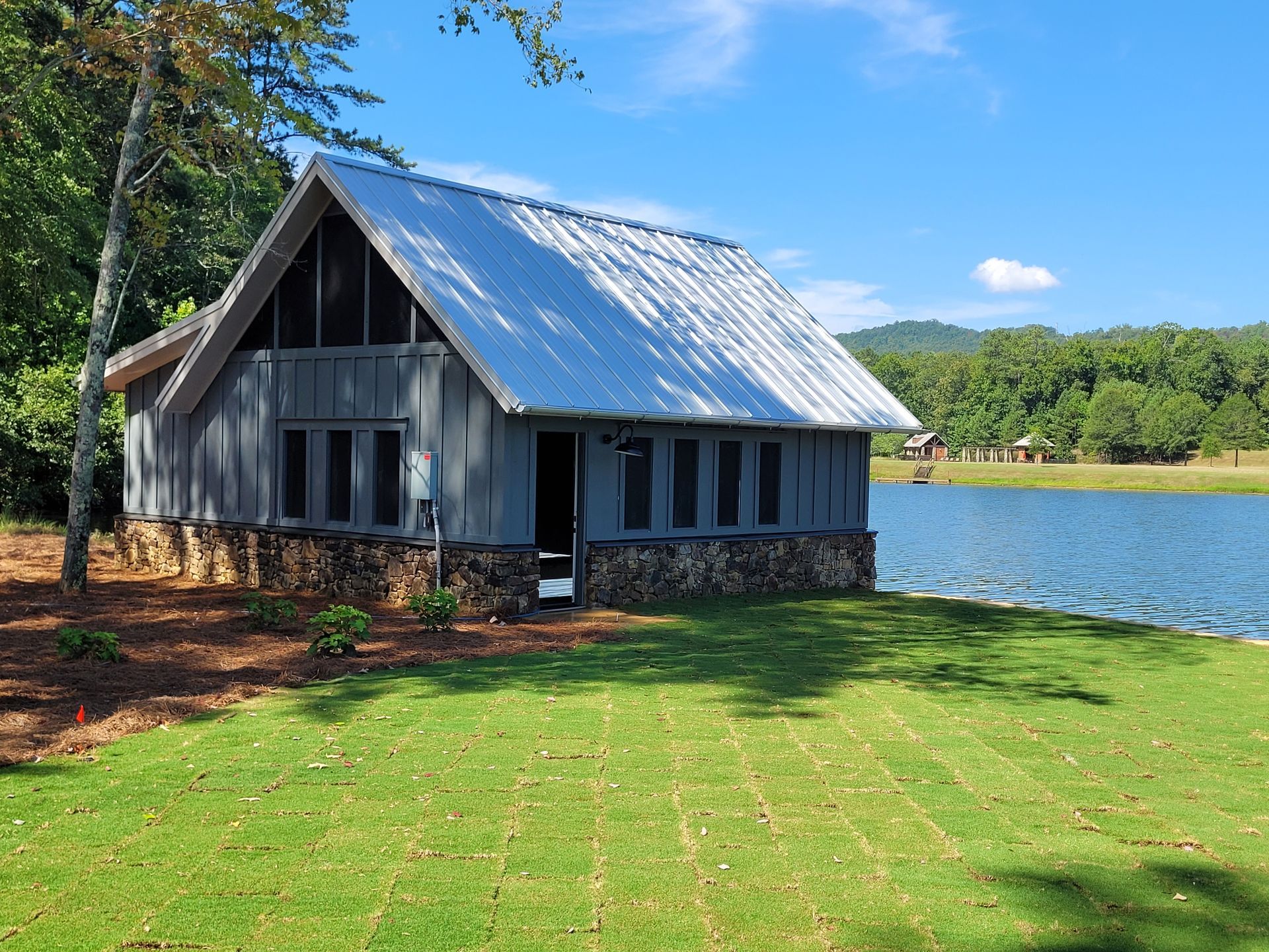 A gray cottage with a metal roof sits next to a lake on a sunny day.