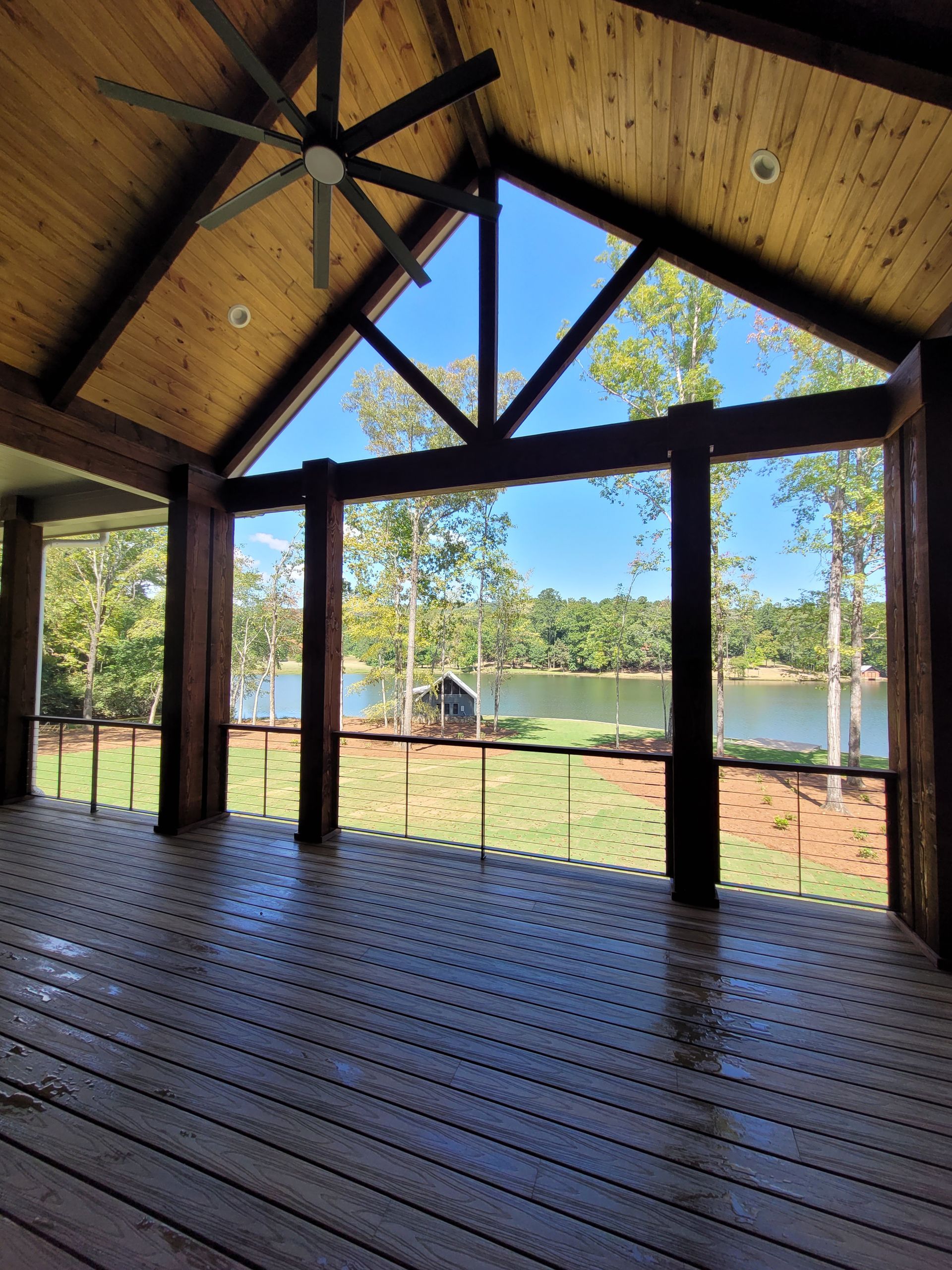 Covered porch overlooking a lake. Wooden beams, ceiling, and deck. Trees and blue sky in the distance.