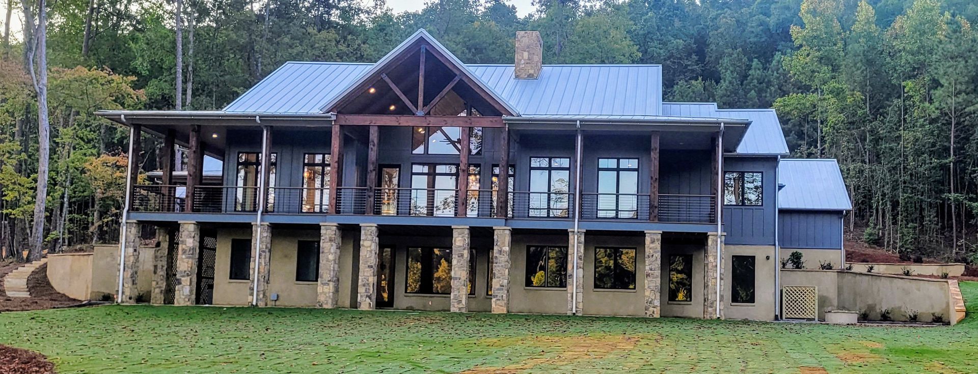 Large, multi-story home with a porch, stone pillars, and a metal roof, set amidst trees.