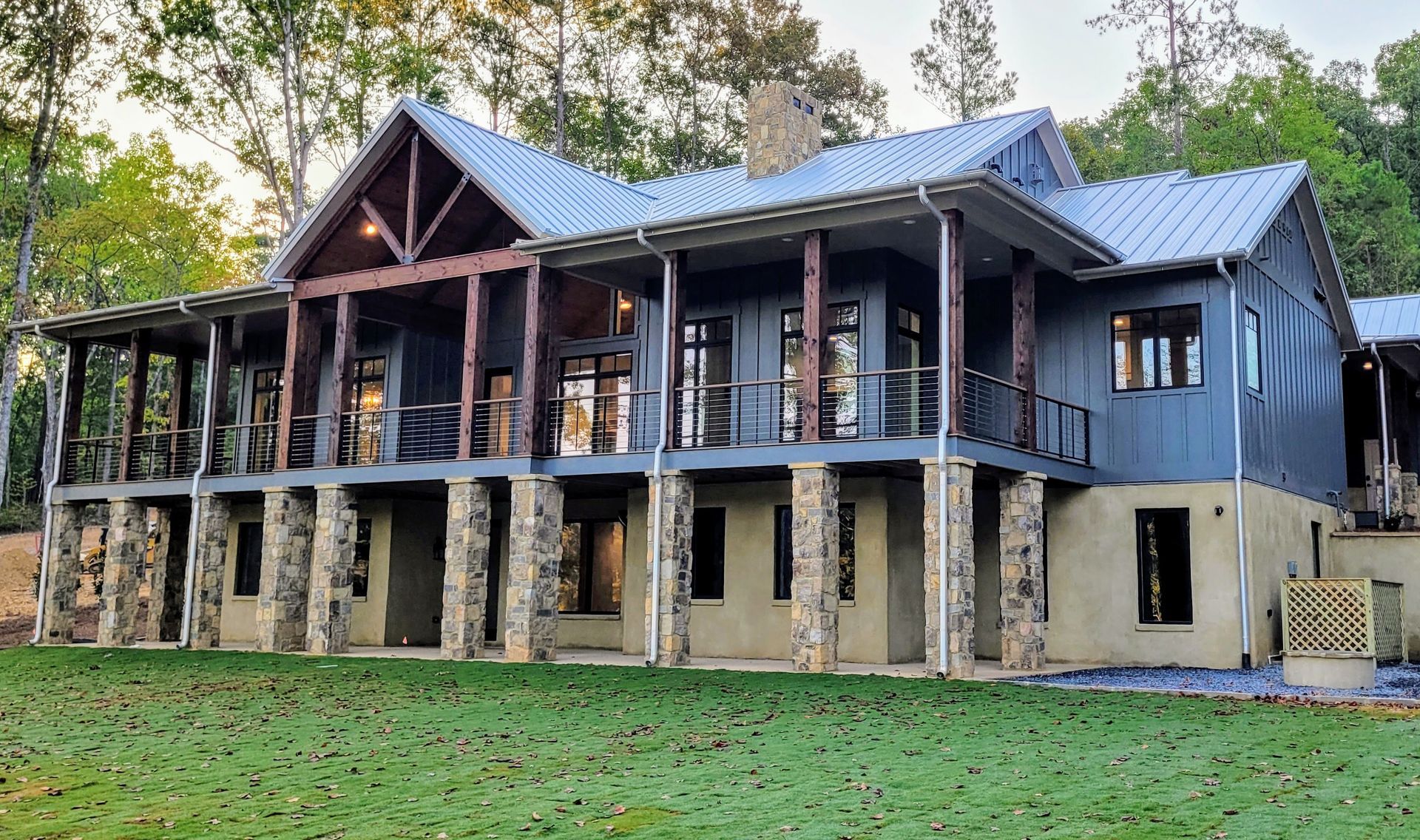 Two-story house with a covered porch, supported by stone columns, nestled in a wooded area. Blue siding, metal roof.