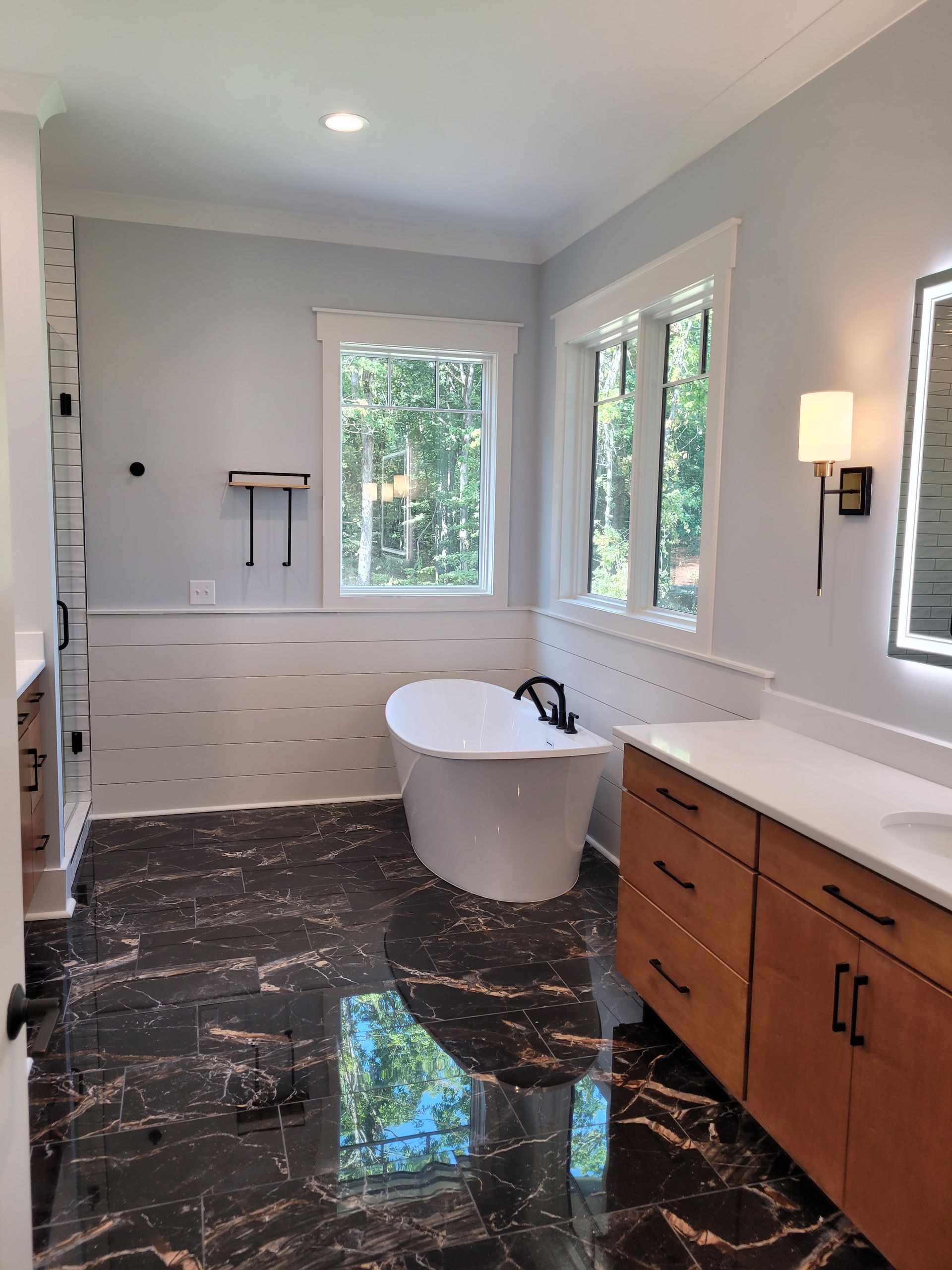 Bathroom with white tub, light blue walls, dark marble floor, wood cabinets, and large window.