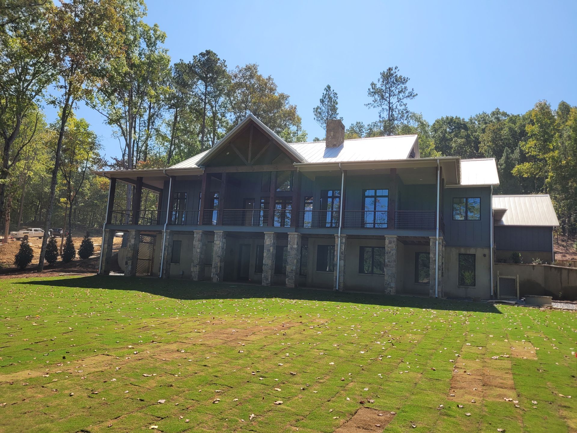 Two-story blue house with stone base and screened porch, surrounded by trees and grassy yard under a blue sky.