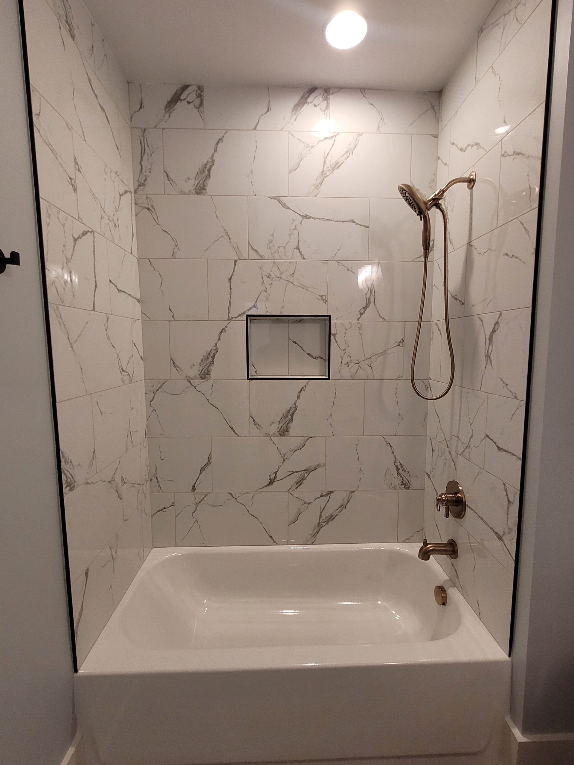 White marble-tiled shower/tub combo with bronze fixtures, built-in shelf, and a single overhead light.