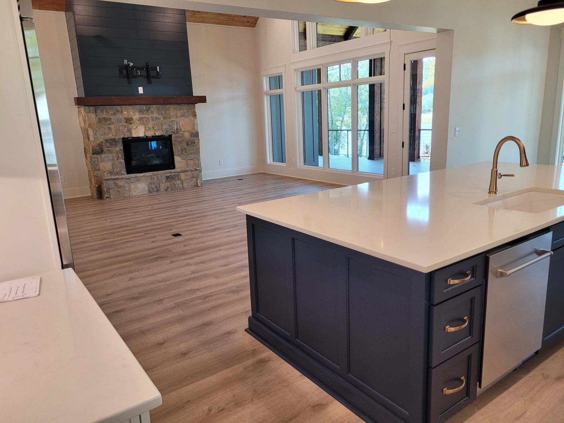 Kitchen island with sink and dishwasher in front of a living room with stone fireplace and large windows.