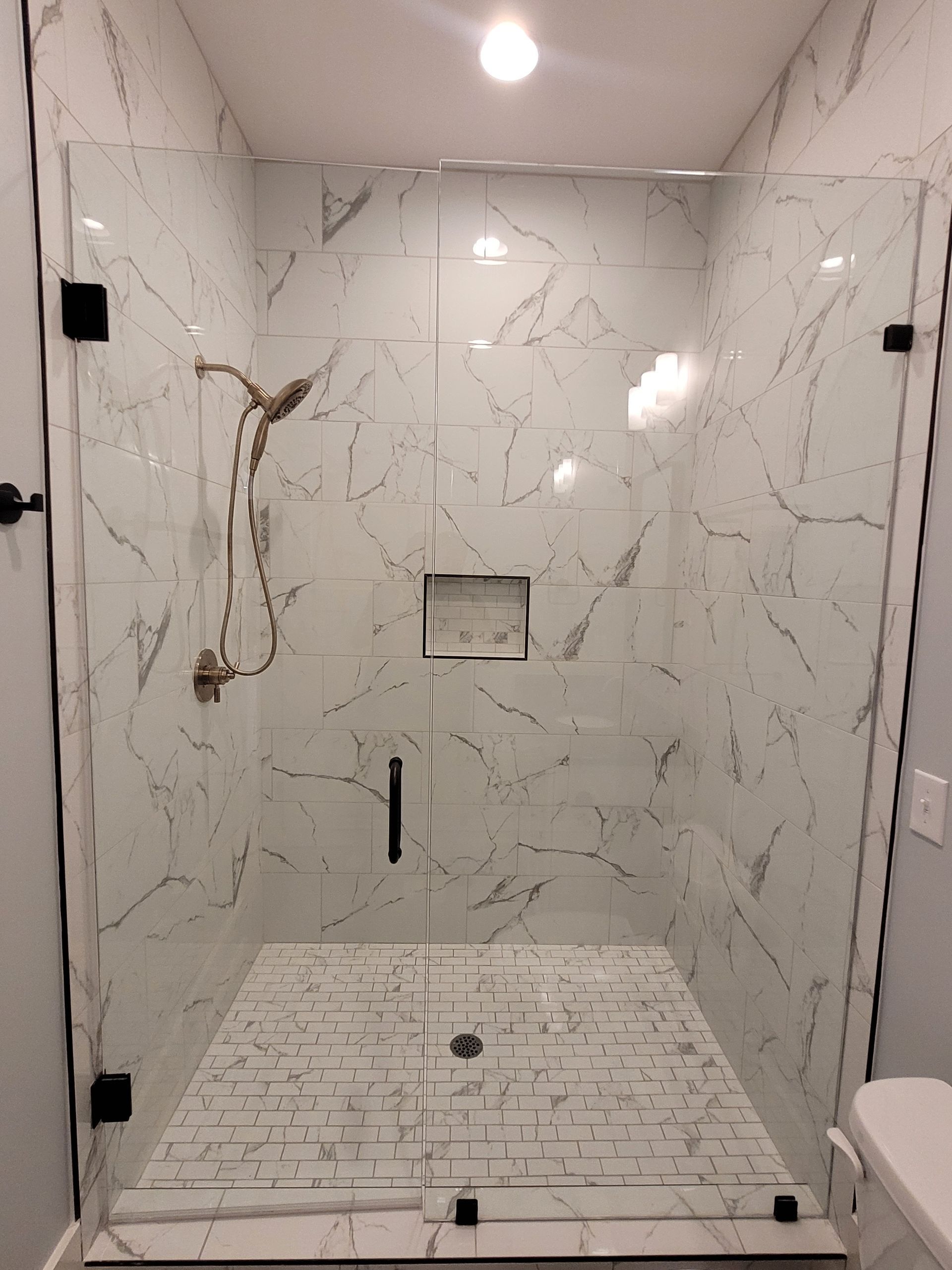 Shower with marble-look tile and glass doors, black hardware, bronze shower head, and recessed shelf.
