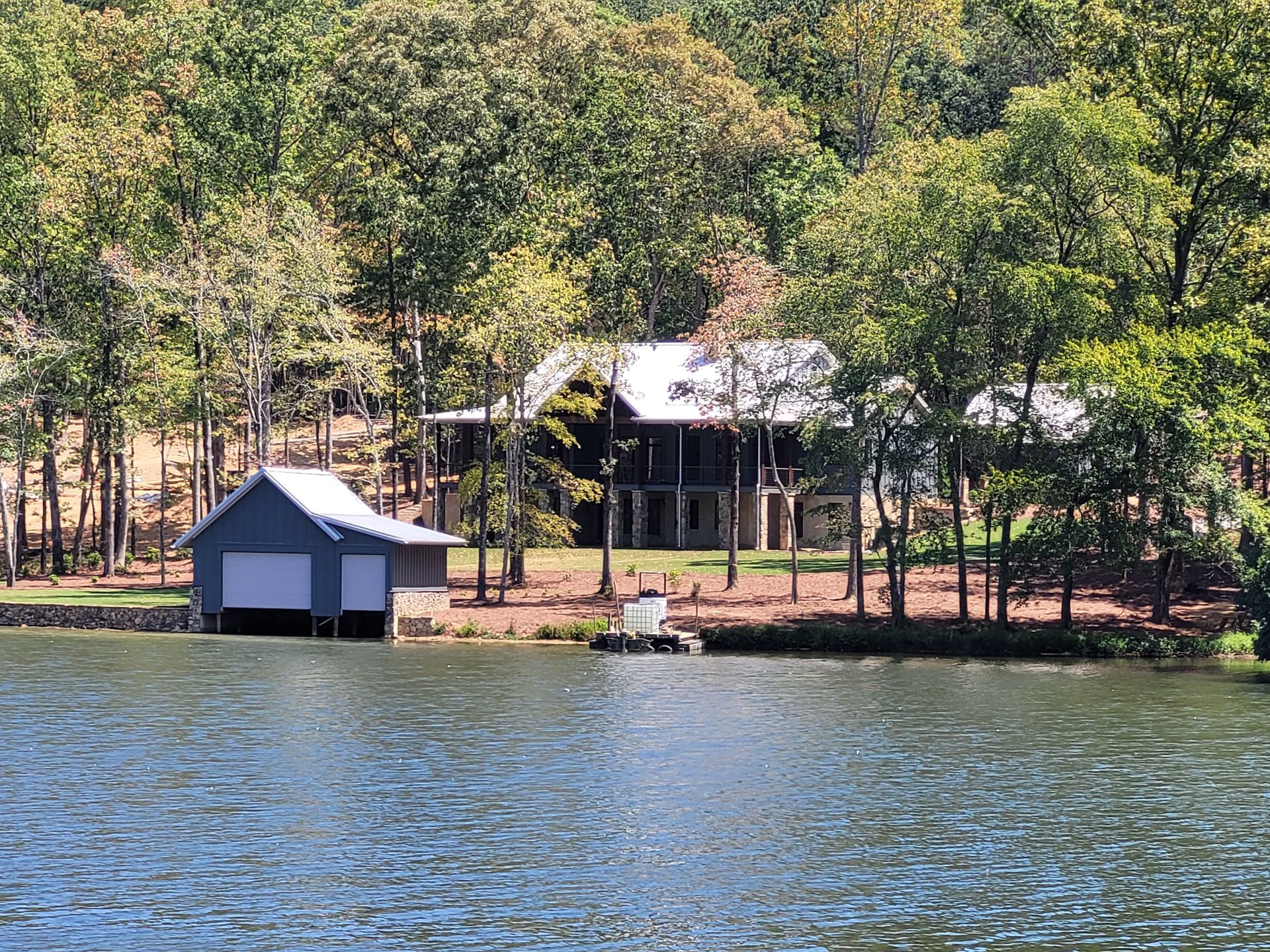 House and boathouse on a lake, surrounded by trees; blue water and sky.