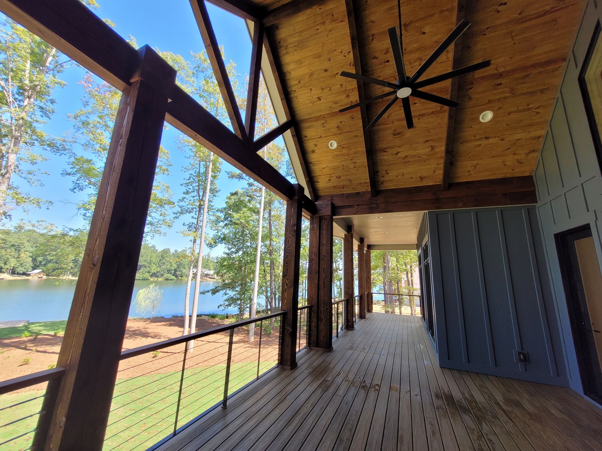 Covered porch overlooking a lake. Dark brown wood beams, light wood ceiling, blue house exterior, green grass and trees.