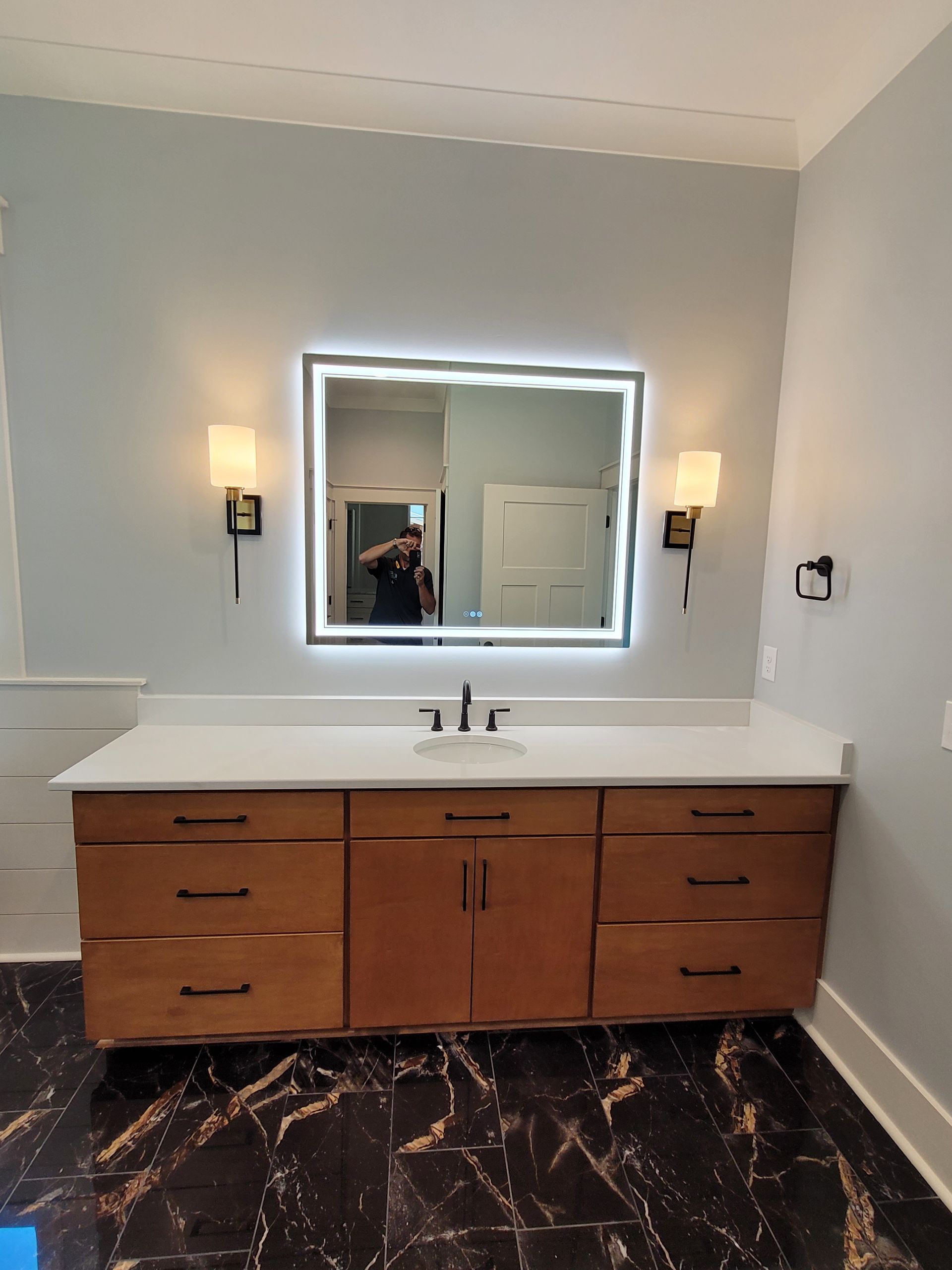Bathroom with a wooden vanity, marble-look floor, backlit mirror, and sconce lights.