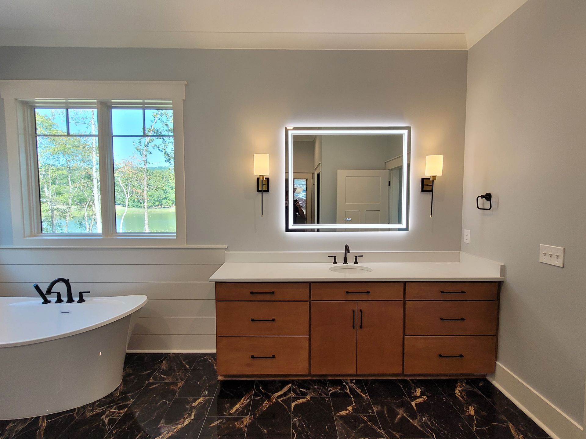 Bathroom with a lighted mirror above a wooden vanity. Bathtub and window are visible.