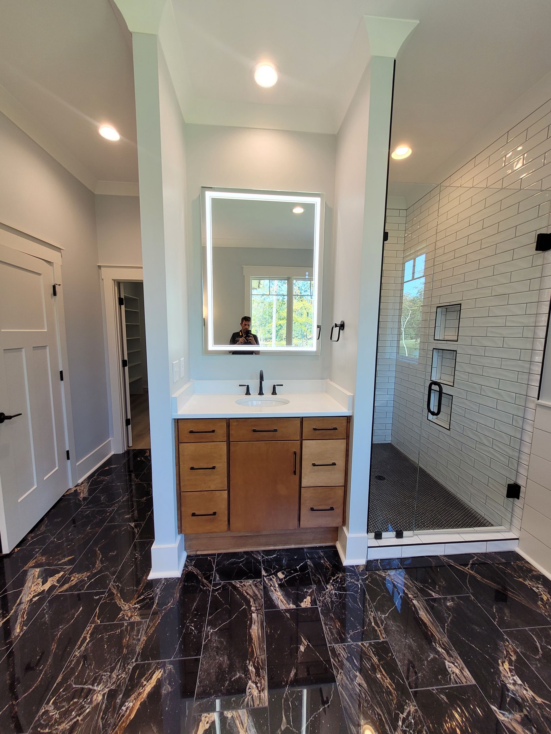 Bathroom with dark patterned floor, wood vanity, lit mirror, and glass shower.