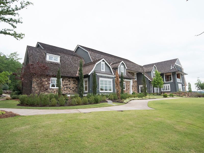 Large, multi-story house with gray siding, stone facade, and brown roof, on a grassy lawn.