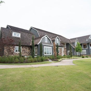 Large house with a stone and gray exterior, brown roof, and a long driveway on a green lawn.