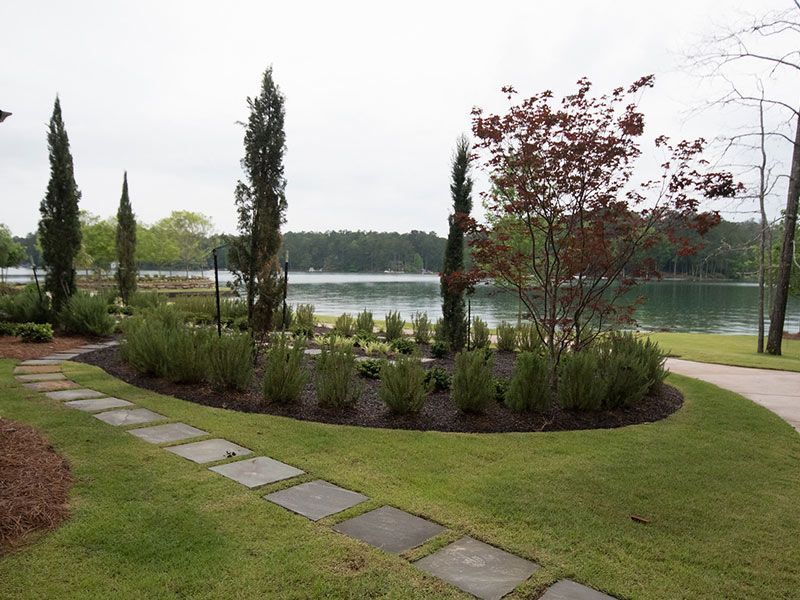 Pathway leading to a lakeside garden with trees, shrubs, and tall, slender evergreens.