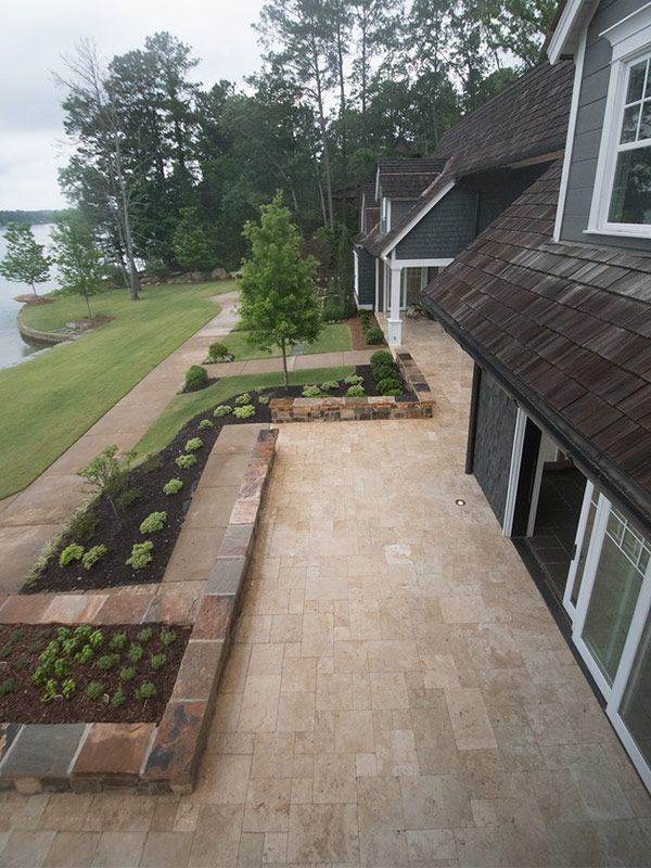 Patio with stone pavers next to a gray house with a dark roof, overlooking a lake.